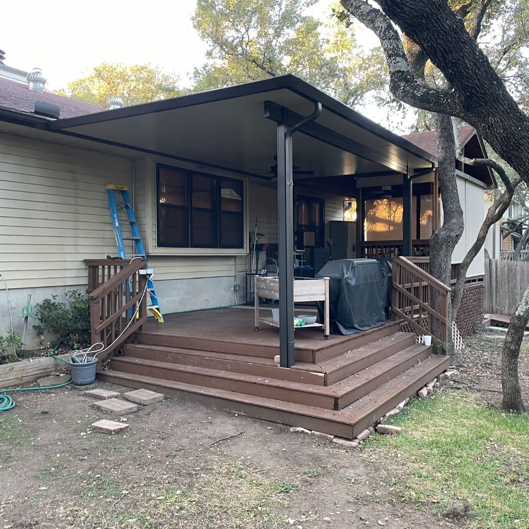 A brown tiered wooden deck attached to a house with a large black roofed patio cover and a blue ladder leaning against it.