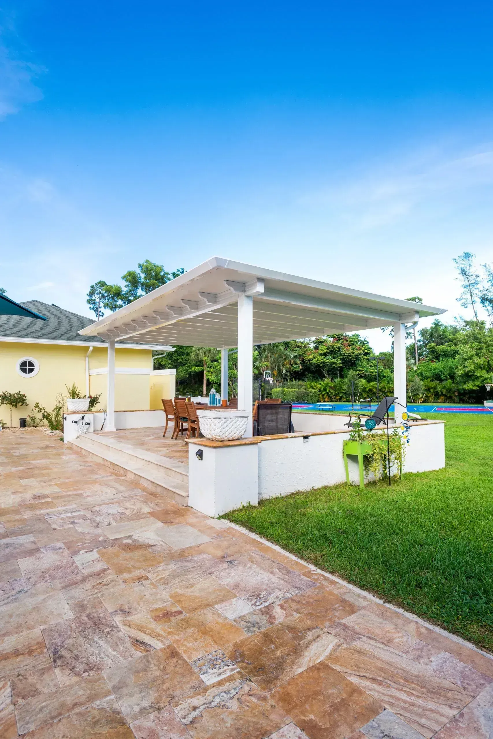 A white pergola shades a stone patio area with seating, set against a green lawn and blue sky.