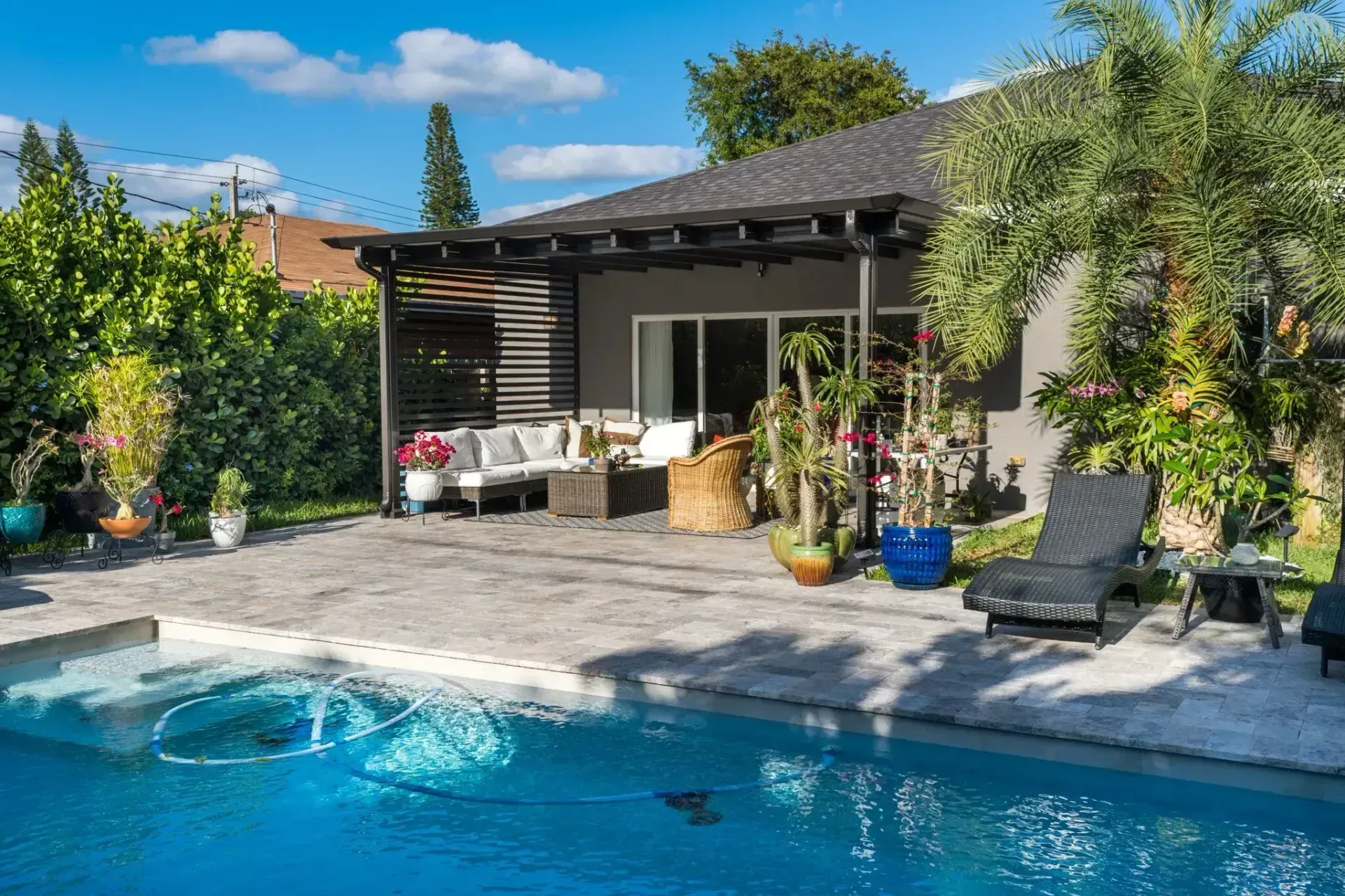 A backyard patio features a pergola, lounge seating, potted plants, and a sun lounger overlooking a blue swimming pool.
