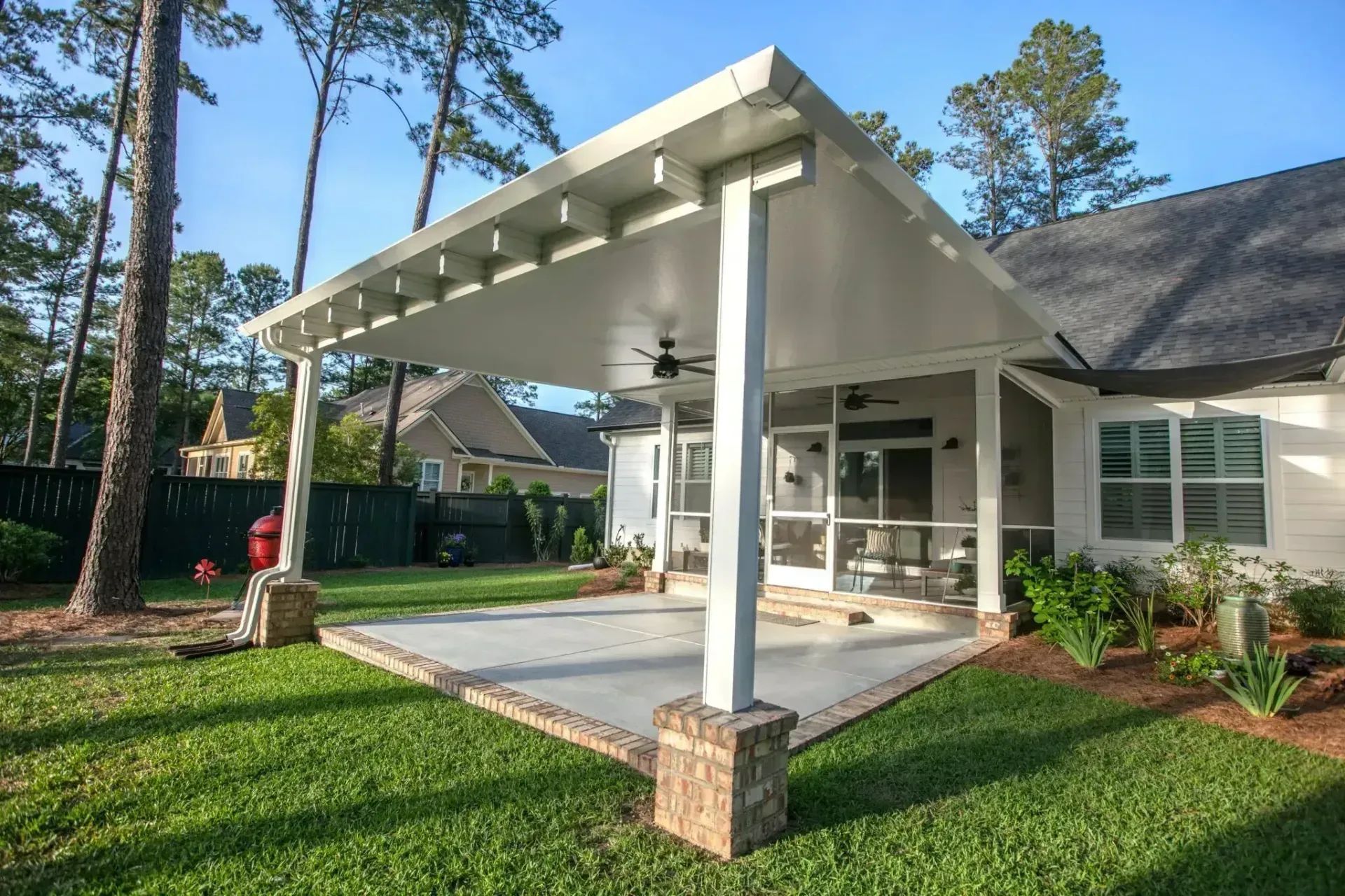Covered patio with white beams, stone columns, and a green lawn beside a house.