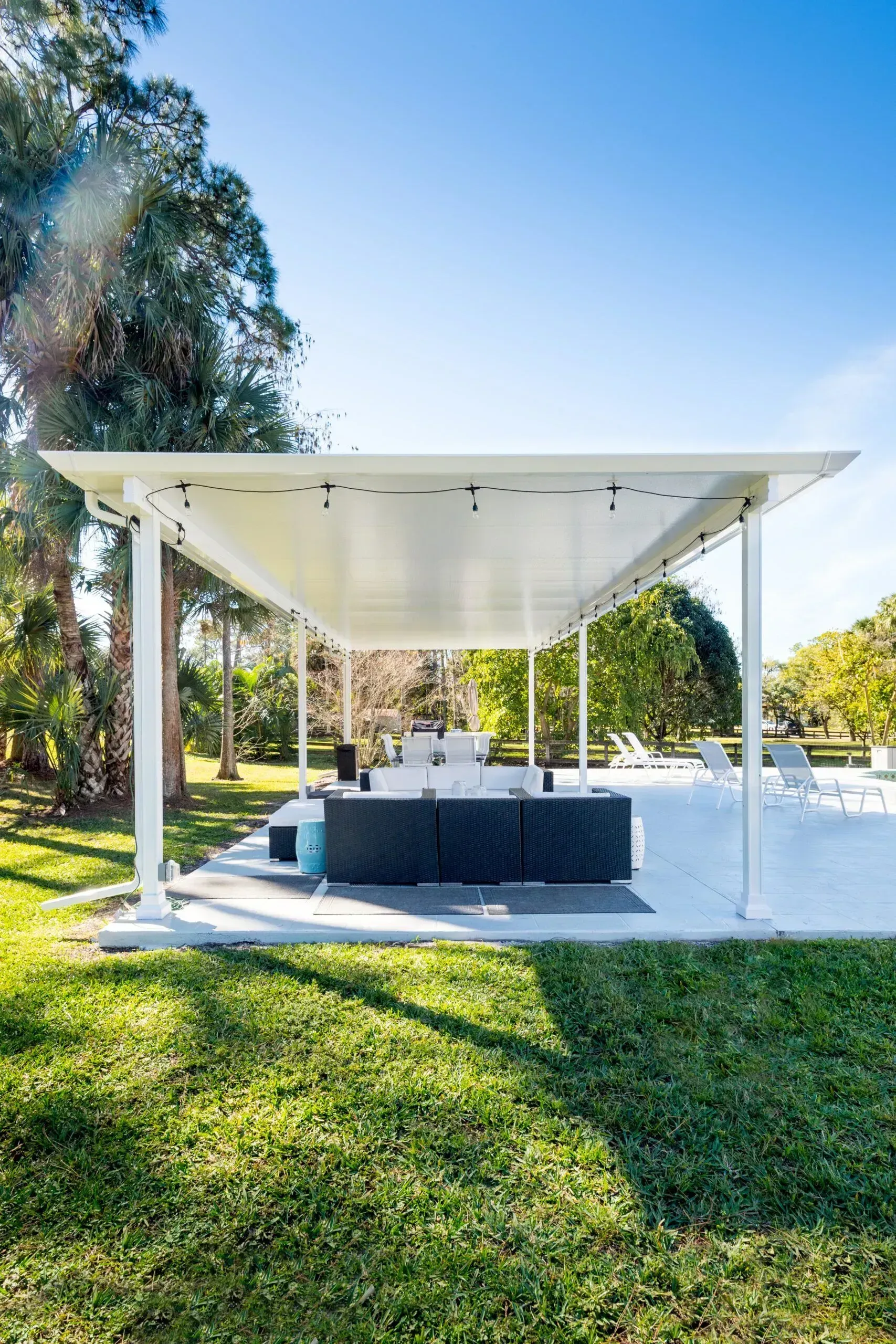 A white patio cover provides shade over a modern outdoor lounge set on a bright, sunny day with green trees in the back.