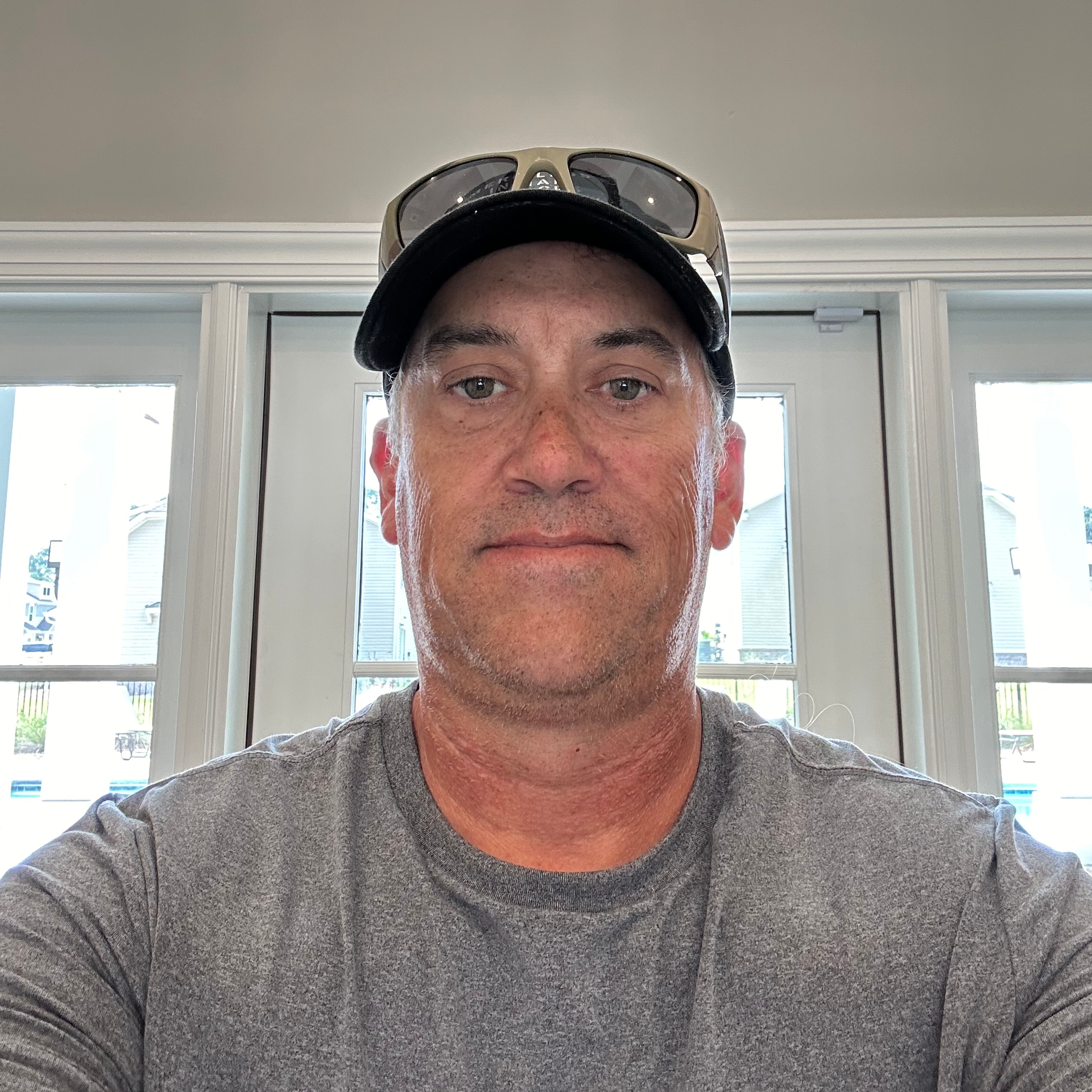 A man wearing a hat and sunglasses is taking a selfie in front of a window.