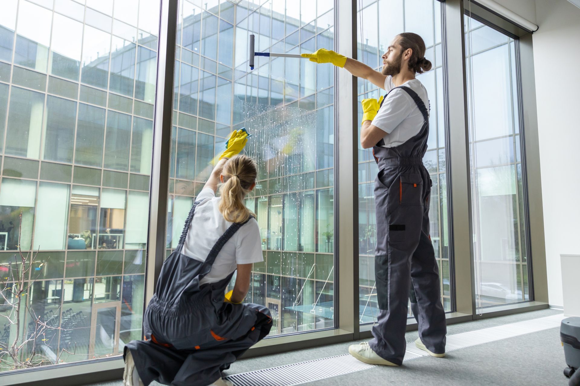 Two window cleaners in gray uniforms clean large office windows with yellow cloths.