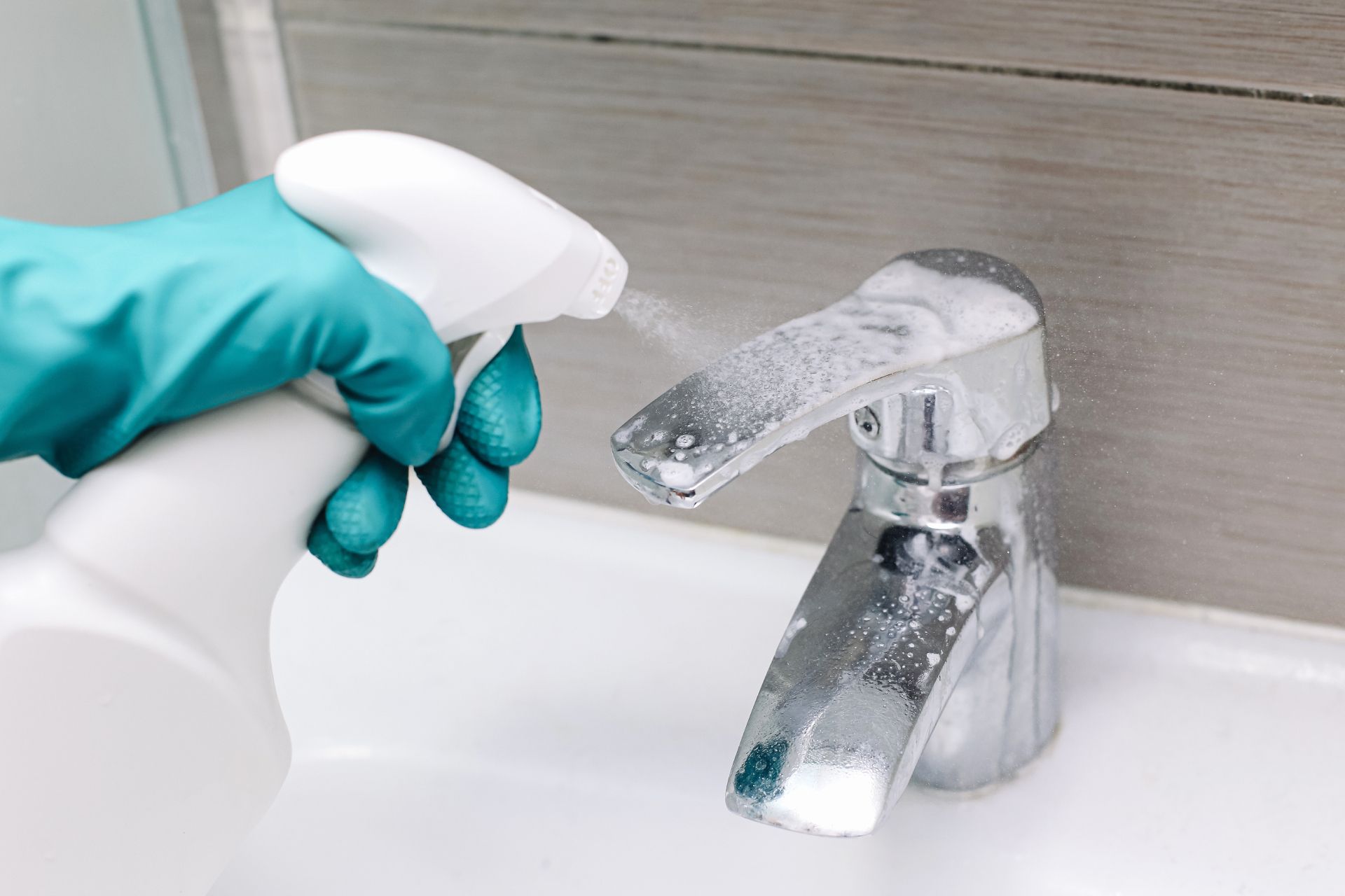 Gloved hand spraying cleaner onto a bathroom faucet and sink