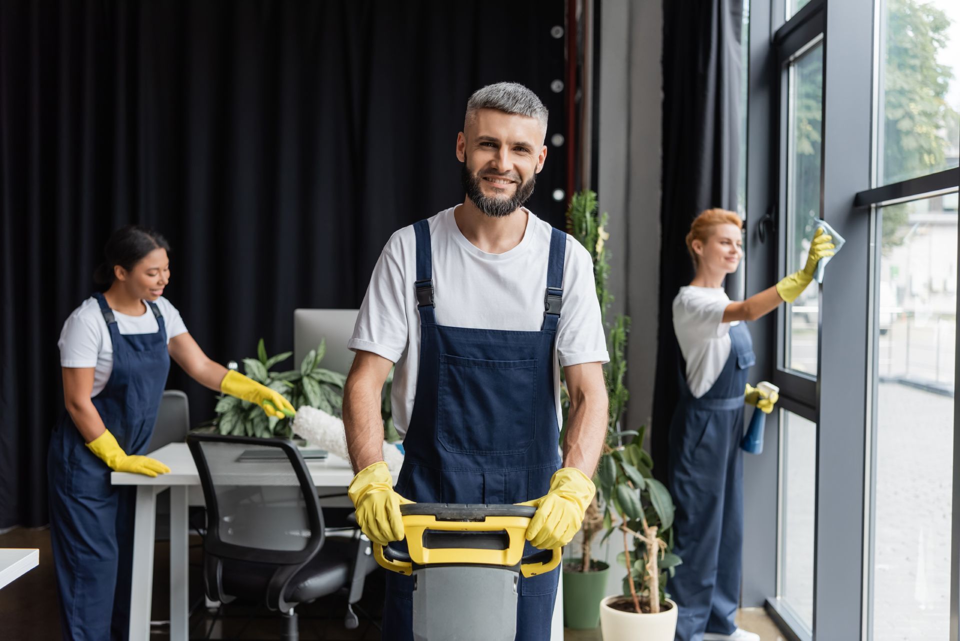 Cleaning crew in blue overalls and yellow gloves washing windows and equipment in an office.