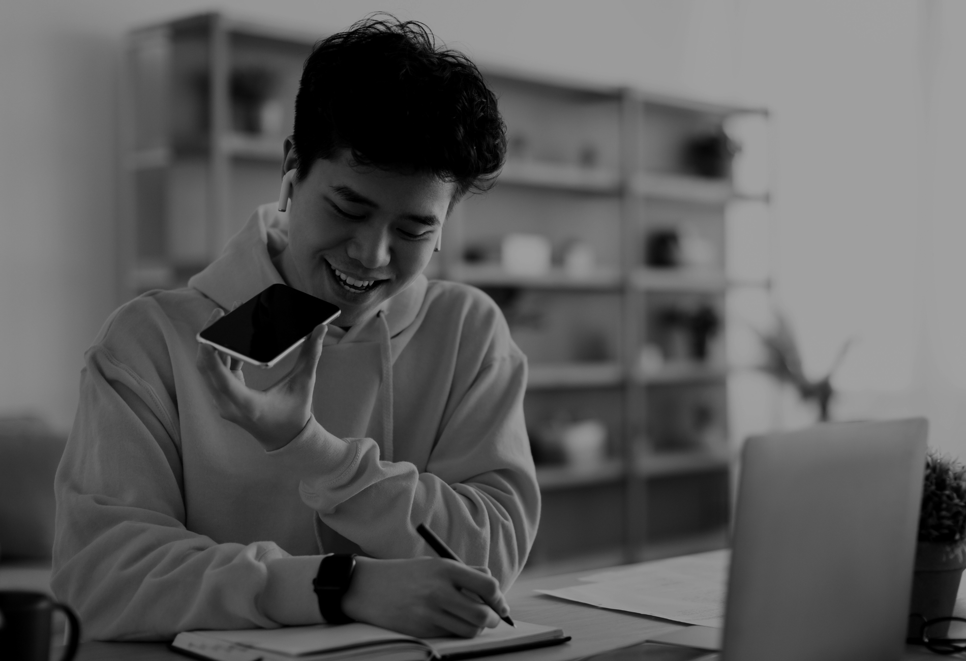 A man is sitting at a desk using a laptop and a cell phone.