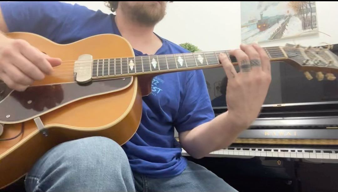 A person in a plaid shirt plays a metallic-colored electric guitar with a pearloid pickguard.
