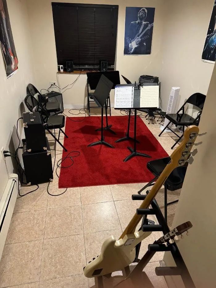 Small music rehearsal room with red rug, music stands, keyboard, guitars, and chairs arranged around them.