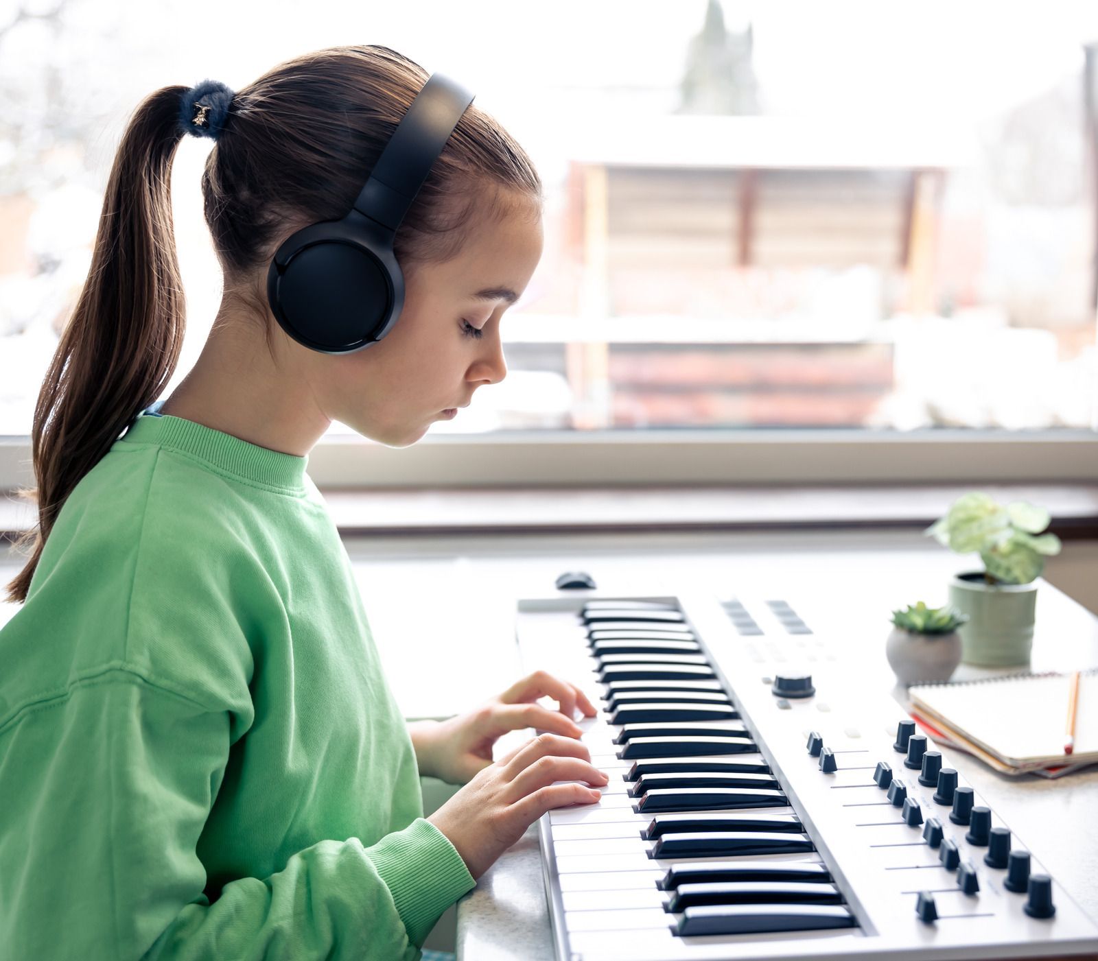 Person in green sweater wearing headphones plays a keyboard by a bright window.