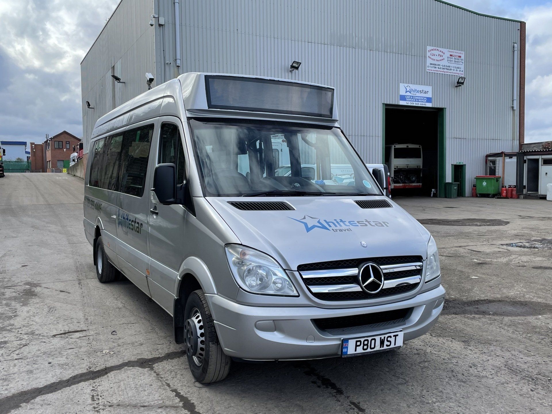 Silver Mercedes-Benz minibus parked outdoors in front of a large industrial building.