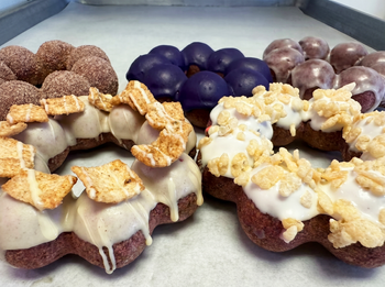 A purple-frosted donut topped with colorful cereal, seen from above with gloved hands at the bottom edges.