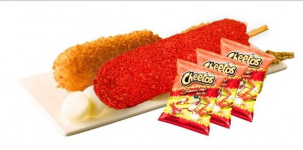 Two breaded, deep-fried corn dogs on a rectangular white plate with lime slices and sausage rounds, on a textured surface.