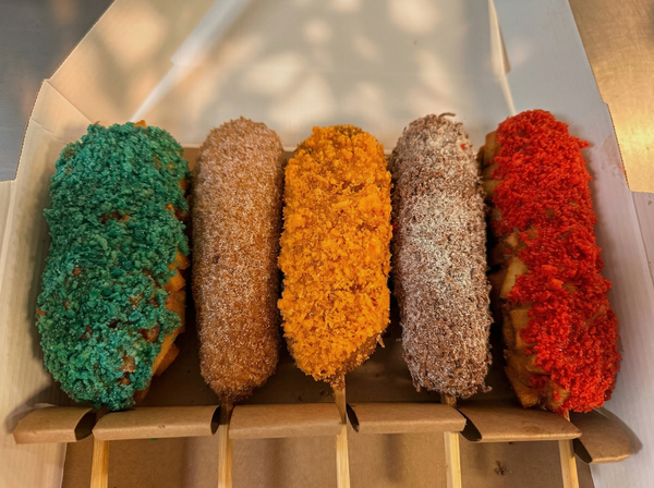 Five Korean-style corn dogs in a box, each with a different colorful, textured coating including green, orange, and red.