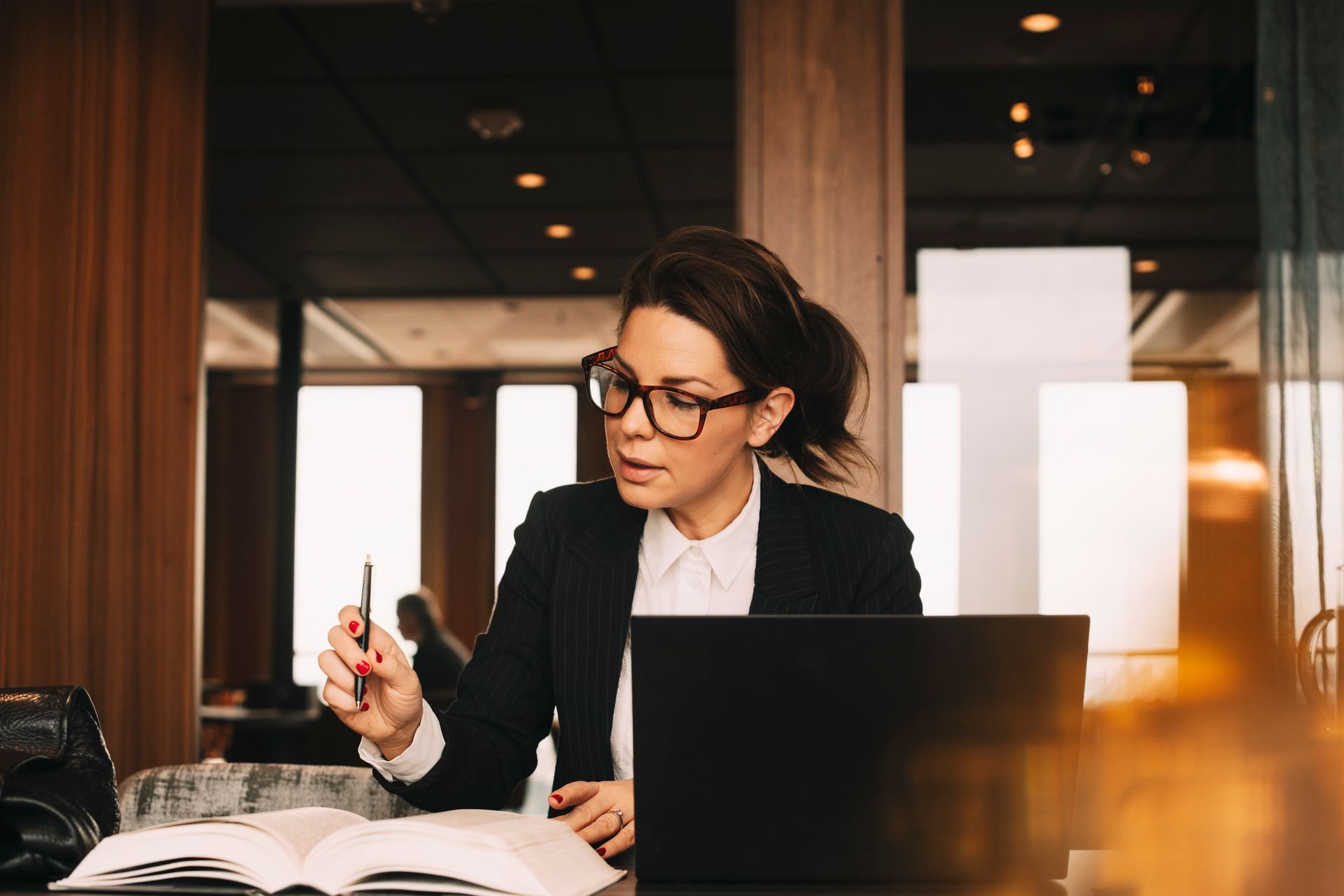 Woman in glasses, suit, working on laptop and reading book in a modern office setting.