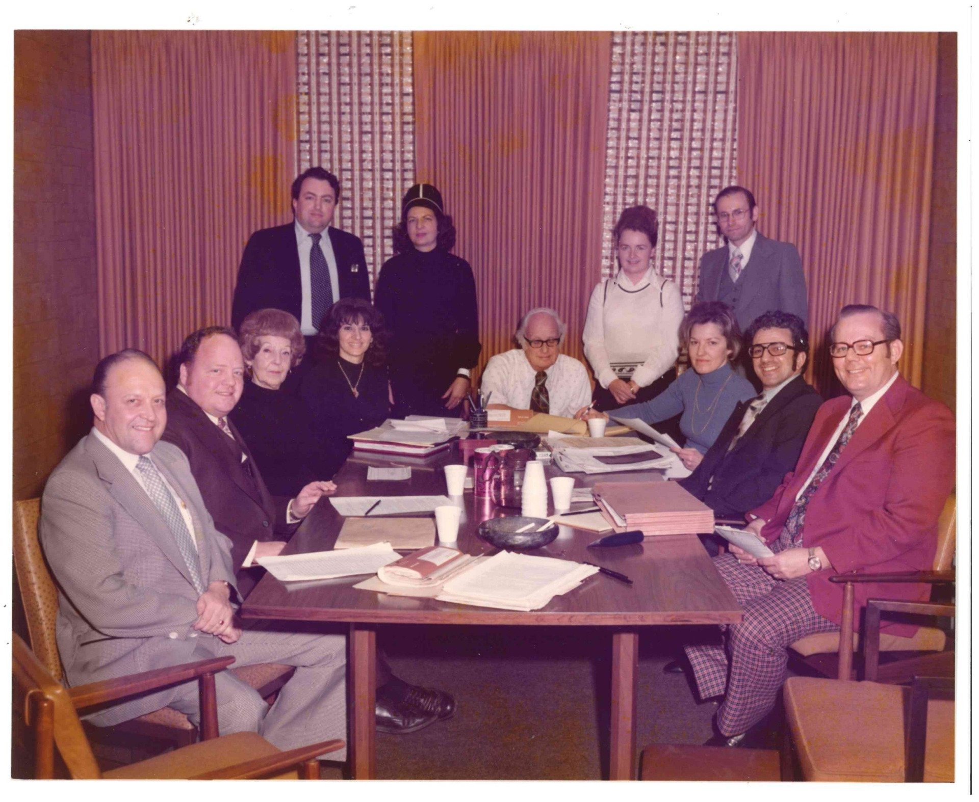 A group of people seated and standing around a conference table in a wood-paneled meeting room.
