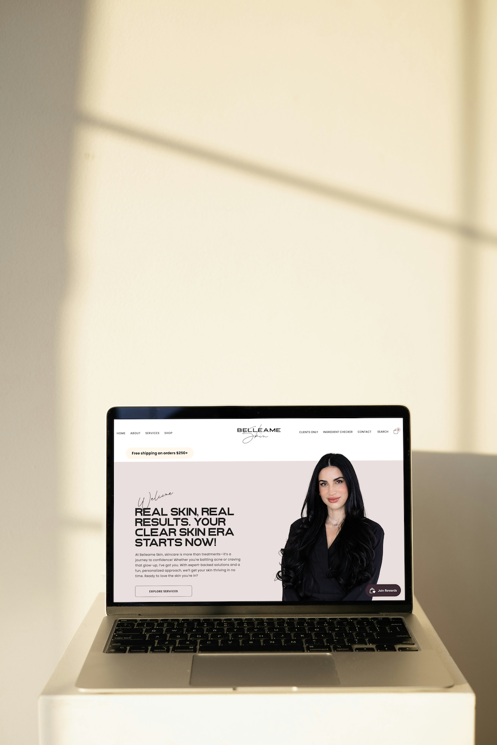 Laptop on a white pedestal displaying a website with a woman and black text against a beige wall.