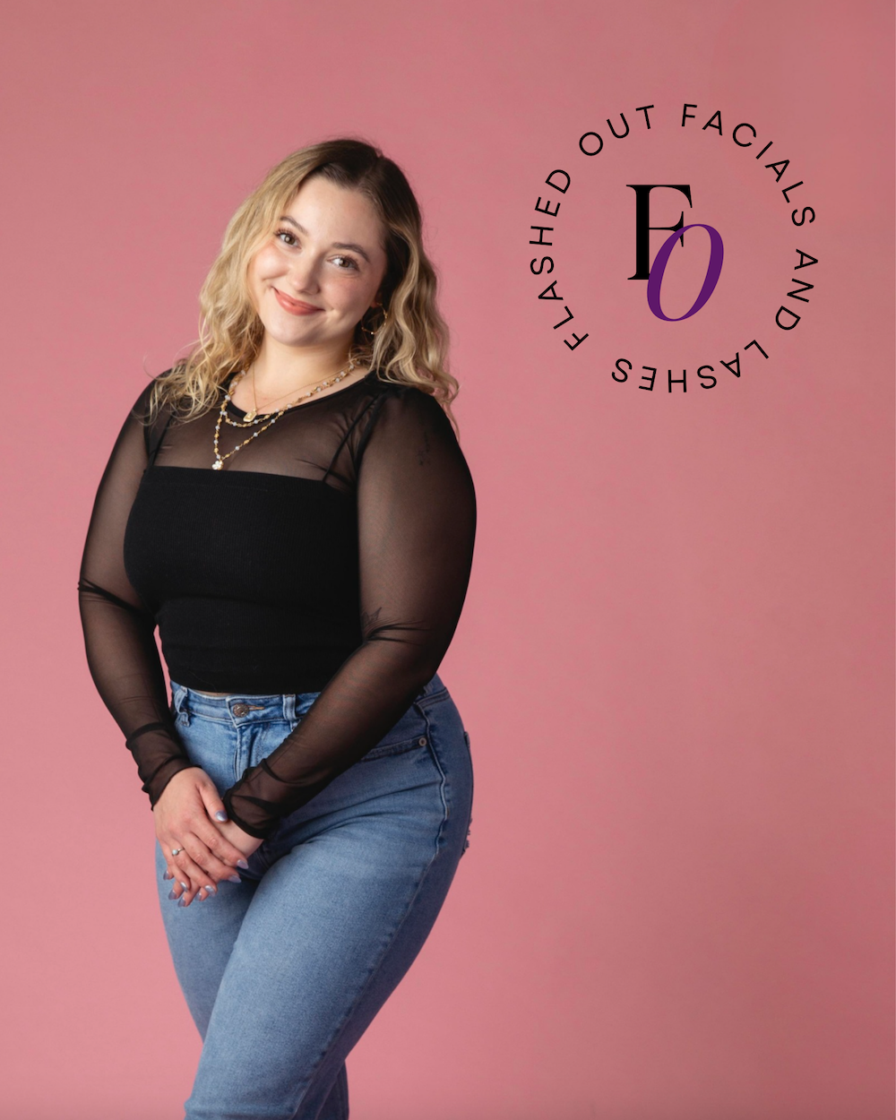 Woman in black top and jeans posing against a pink studio backdrop with a circular logo.