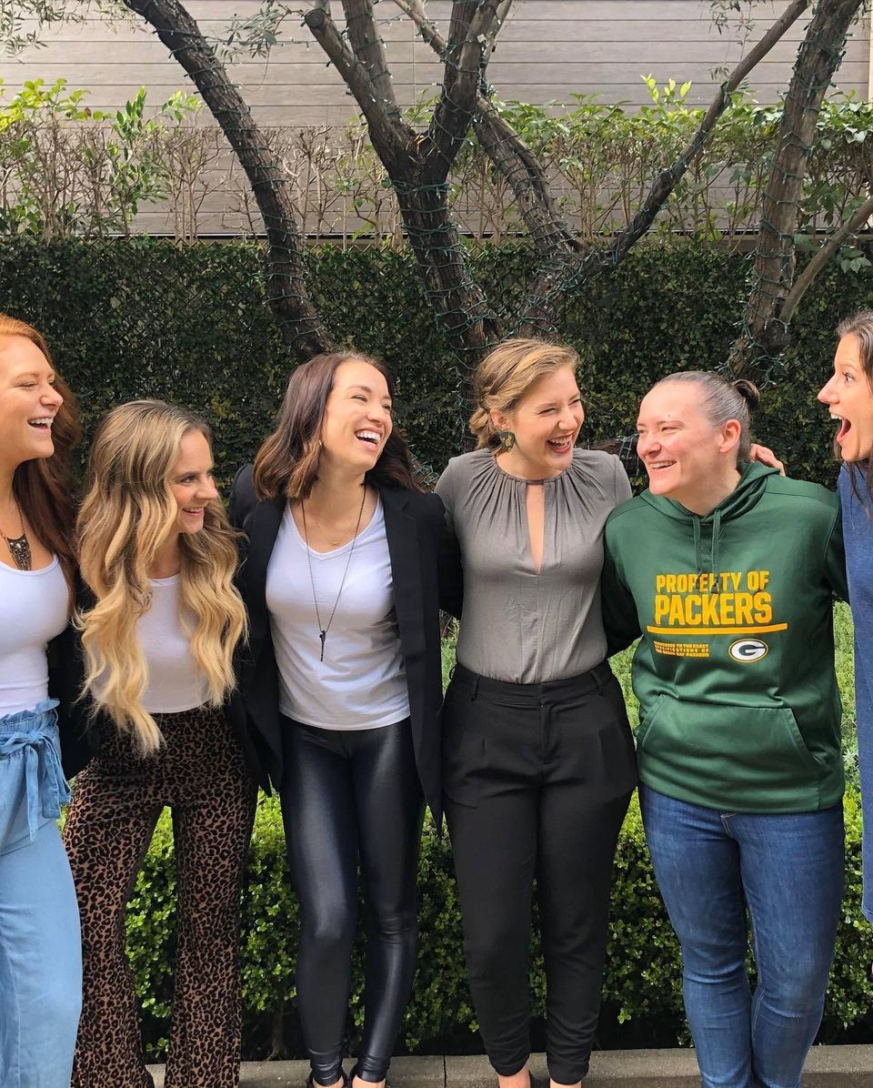 Five women standing arm-in-arm outdoors, laughing in front of a leafy wall.
