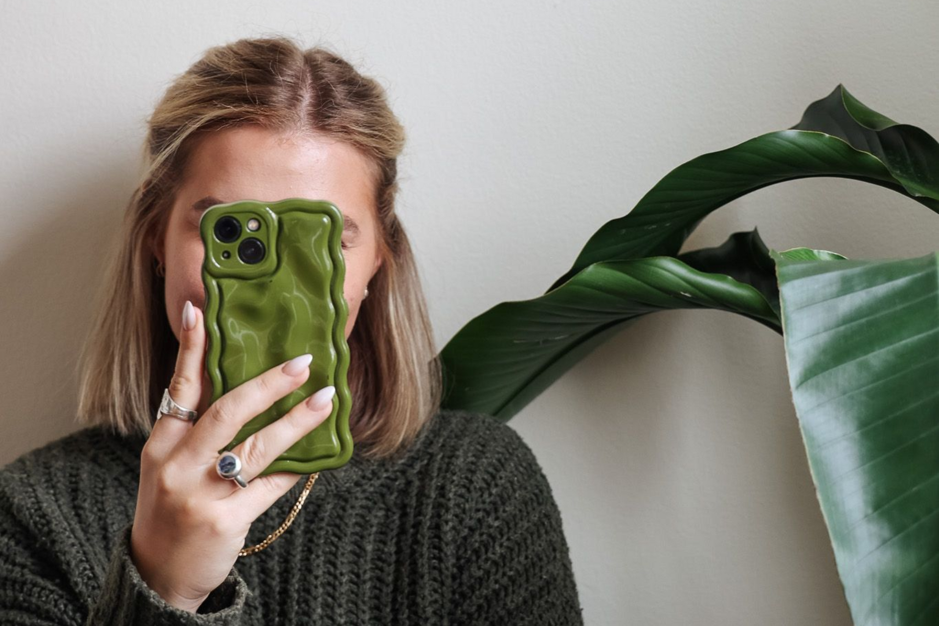 Person taking a mirror selfie in a dark green sweater, with a large leafy plant beside them.
