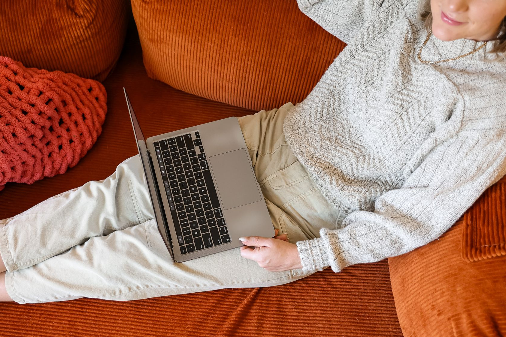 Person reclining on an orange couch using a laptop, wearing a light gray sweater and white pants.