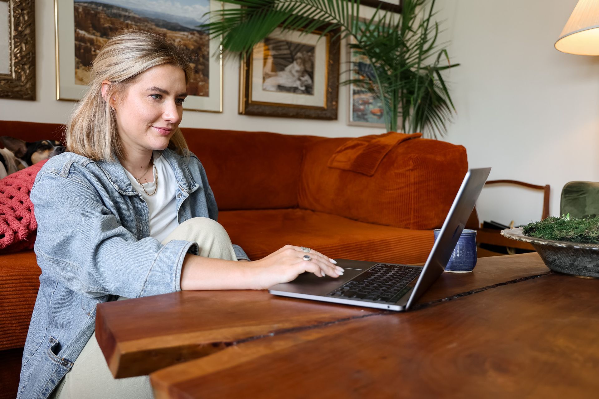 Woman using a laptop at a wooden table in a cozy living room with framed wall art and plants.