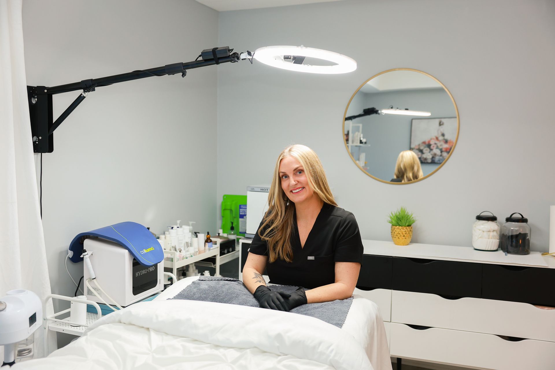 Woman in a spa treatment room seated on a bed under a ring light, with a mirror and cabinet behind her.