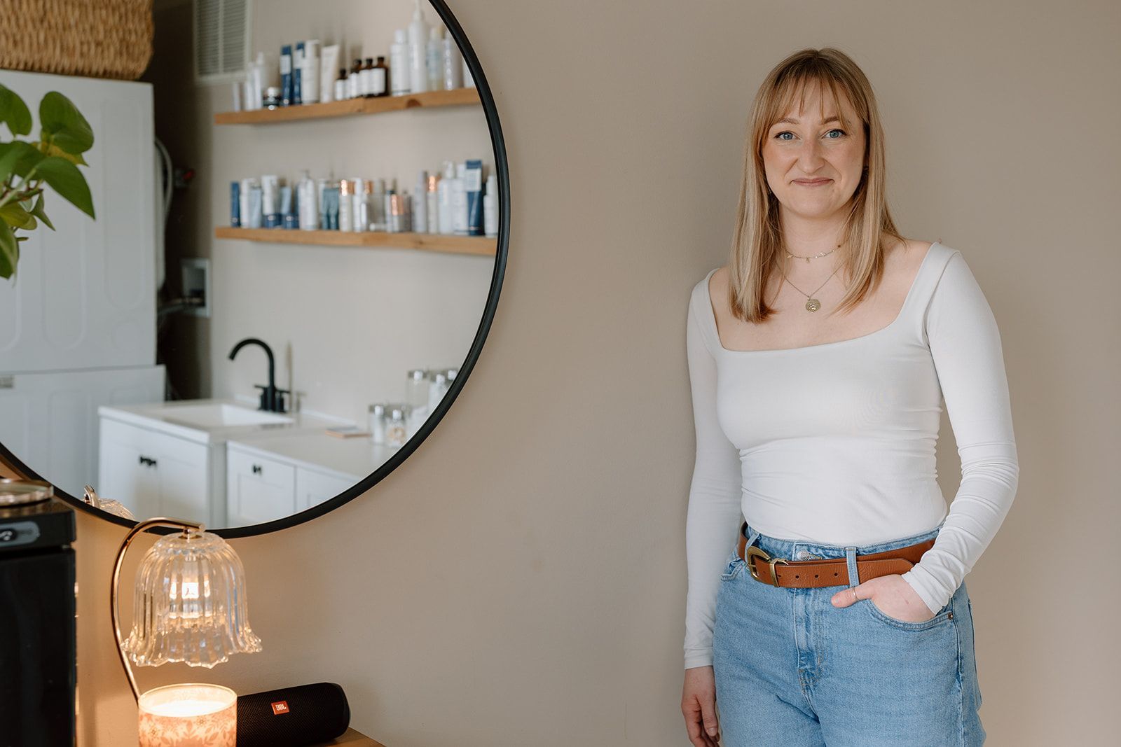 Woman standing in a cozy room beside a round mirror, shelves, and a glowing lamp.