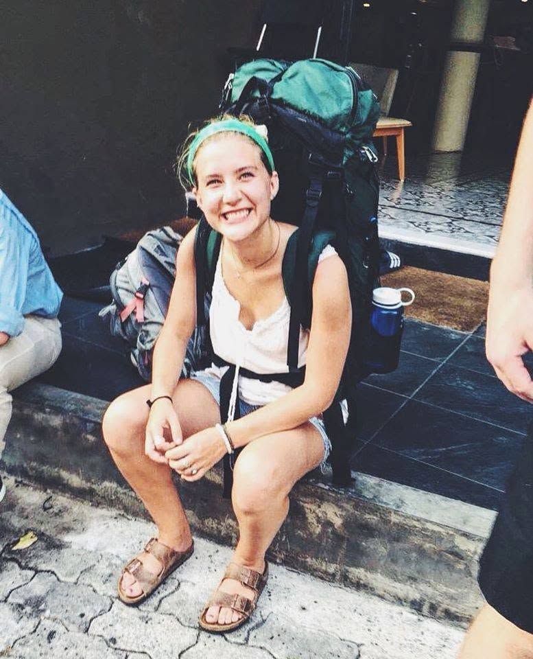 Smiling hiker crouches on stone steps with a large backpack and sandals.