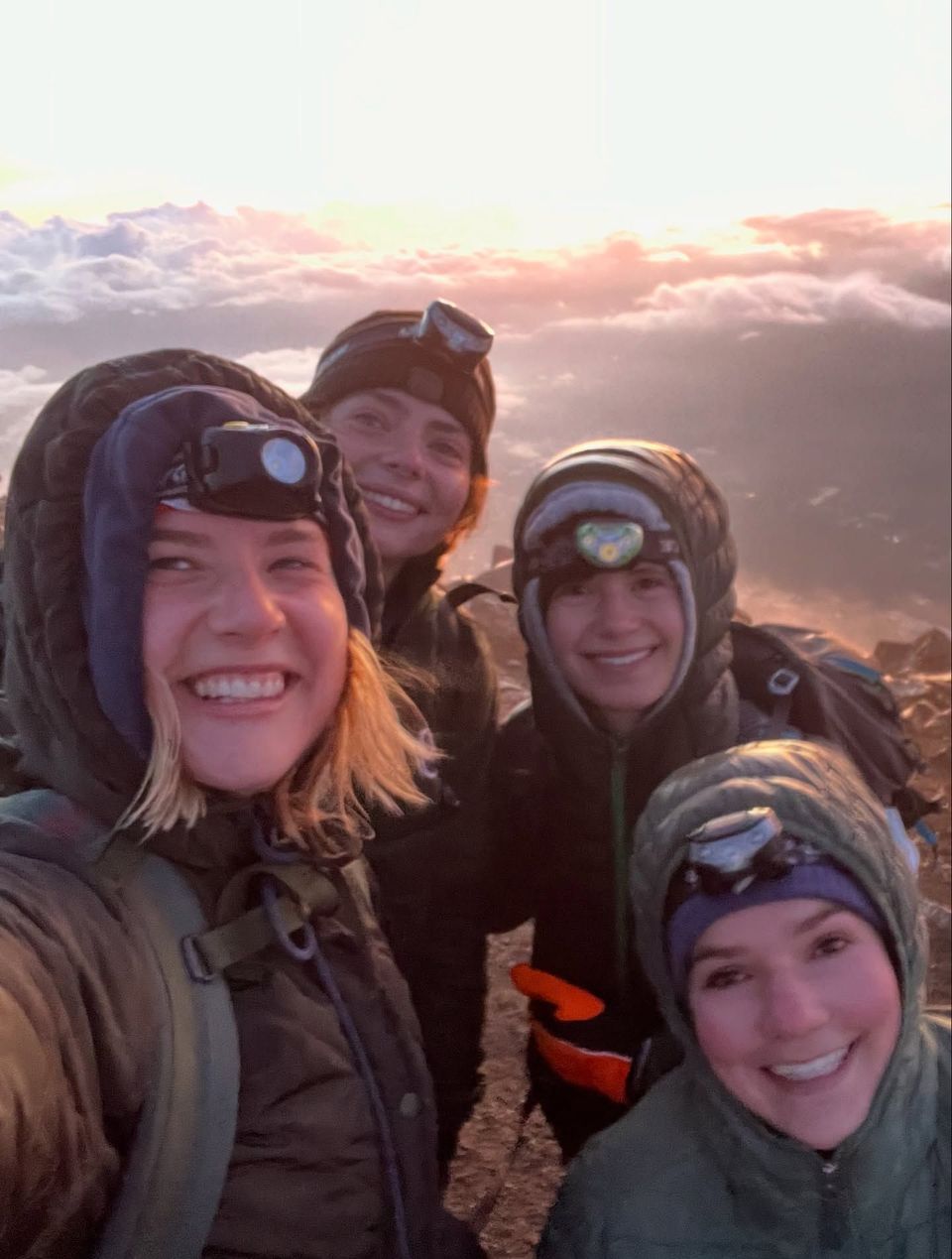 Four friends in winter hats smile for a sunrise selfie on a mountain summit.