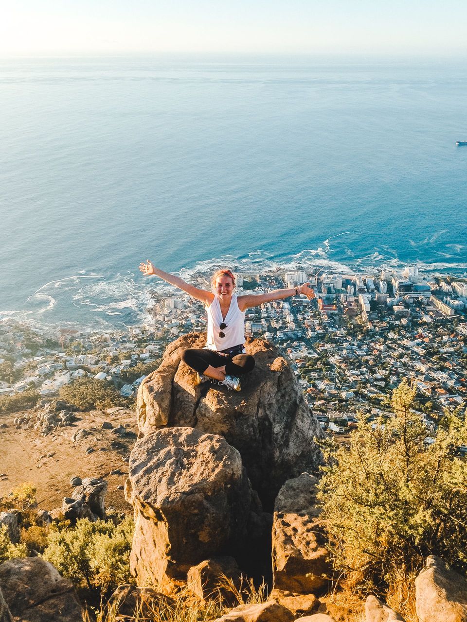 Person standing on a rocky cliff above a seaside town, arms outstretched toward the ocean