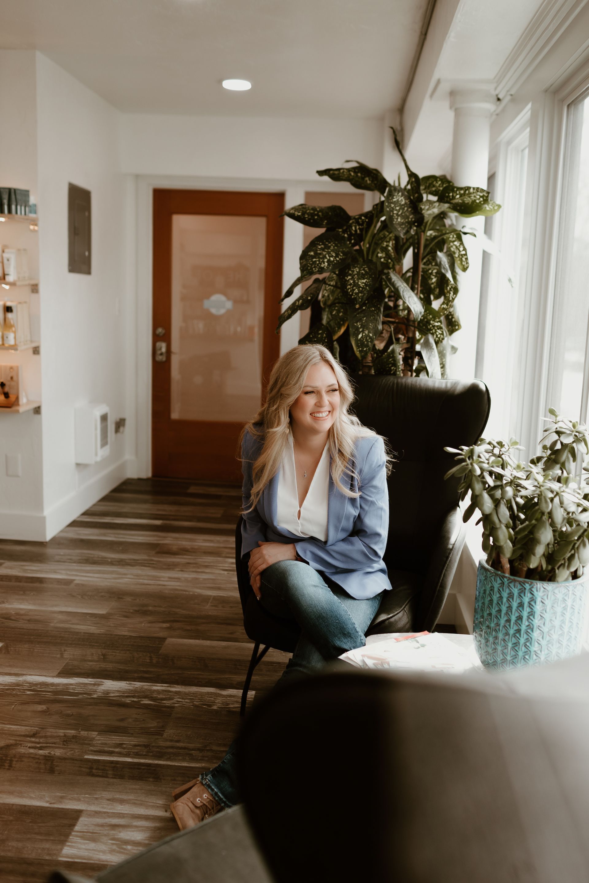 Woman seated in a bright hallway beside large potted plants and wood floors
