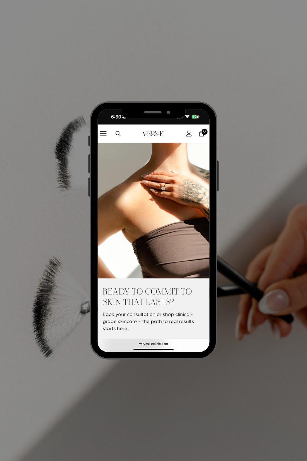 A phone displays a fashion ad while a hand writes beside it on a gray surface.