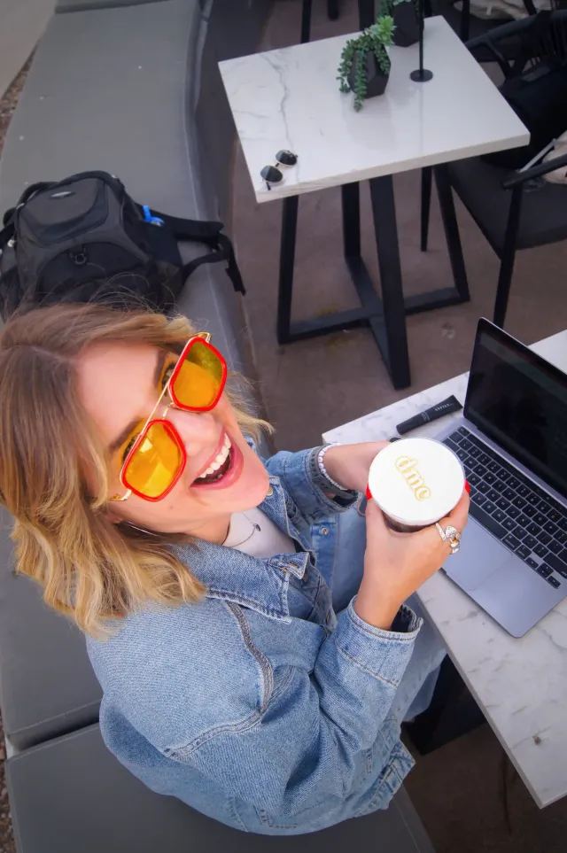Smiling person in orange sunglasses holding a coffee beside a laptop at an outdoor table
