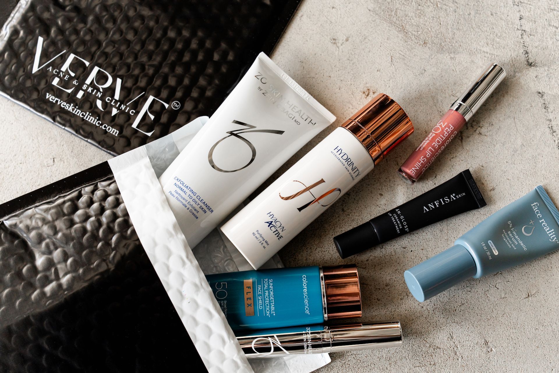 Assorted makeup and skincare products arranged on a textured surface with a black pouch and packaging.