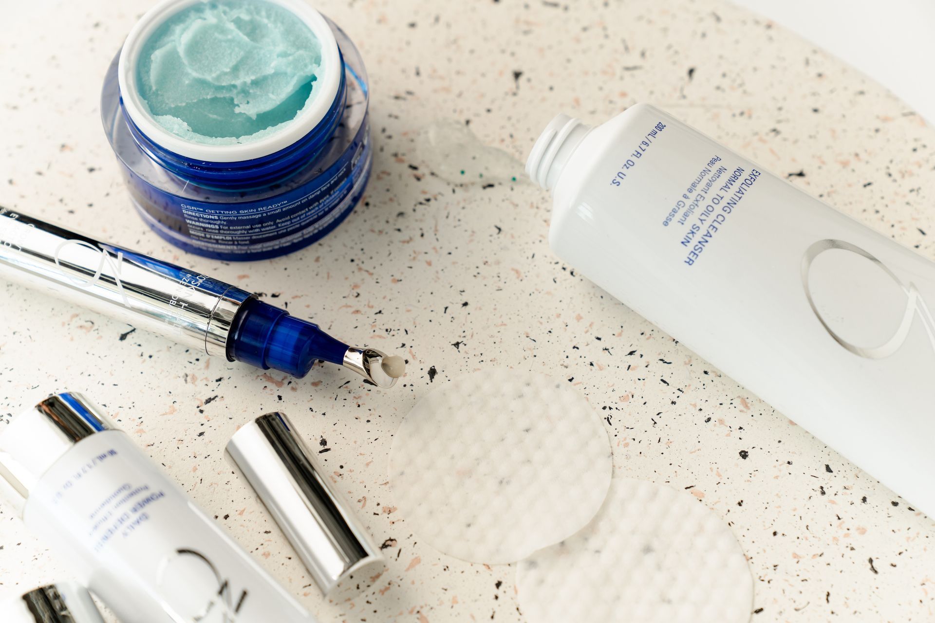 Skincare products on a speckled countertop, including cream jar, serum dropper, and tube.