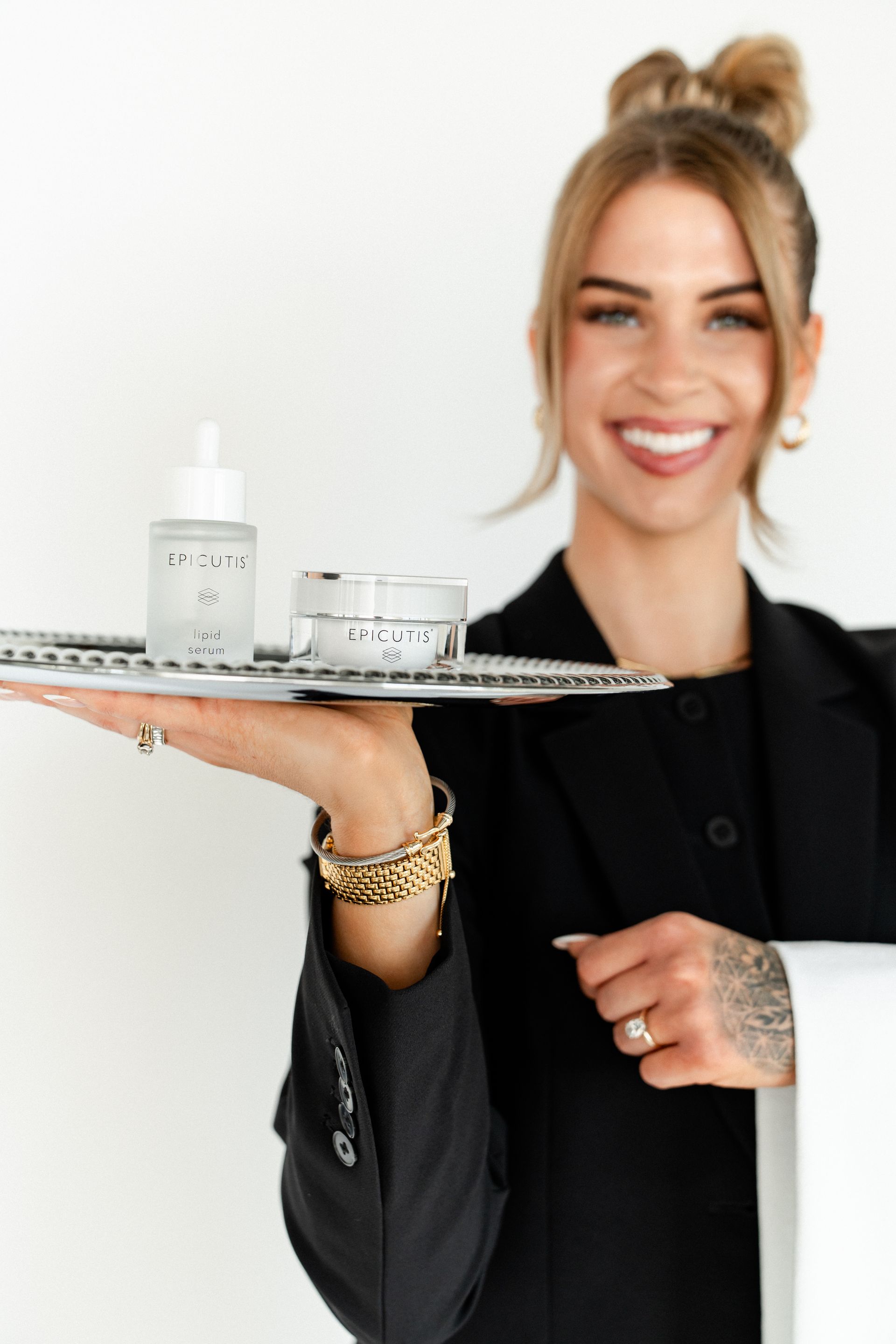 Smiling woman in black blazer holding a tray with white skincare products and jars