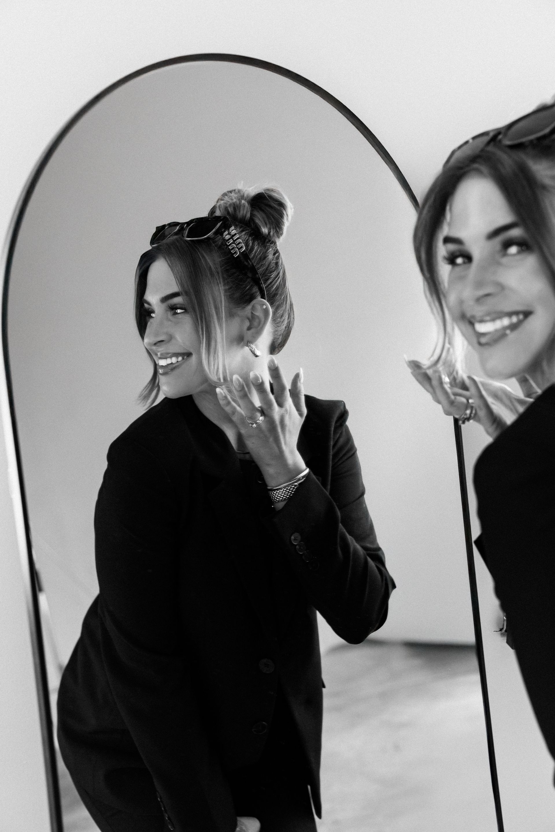 Woman smiling in a mirror, posing in a dark outfit beside a large oval mirror.