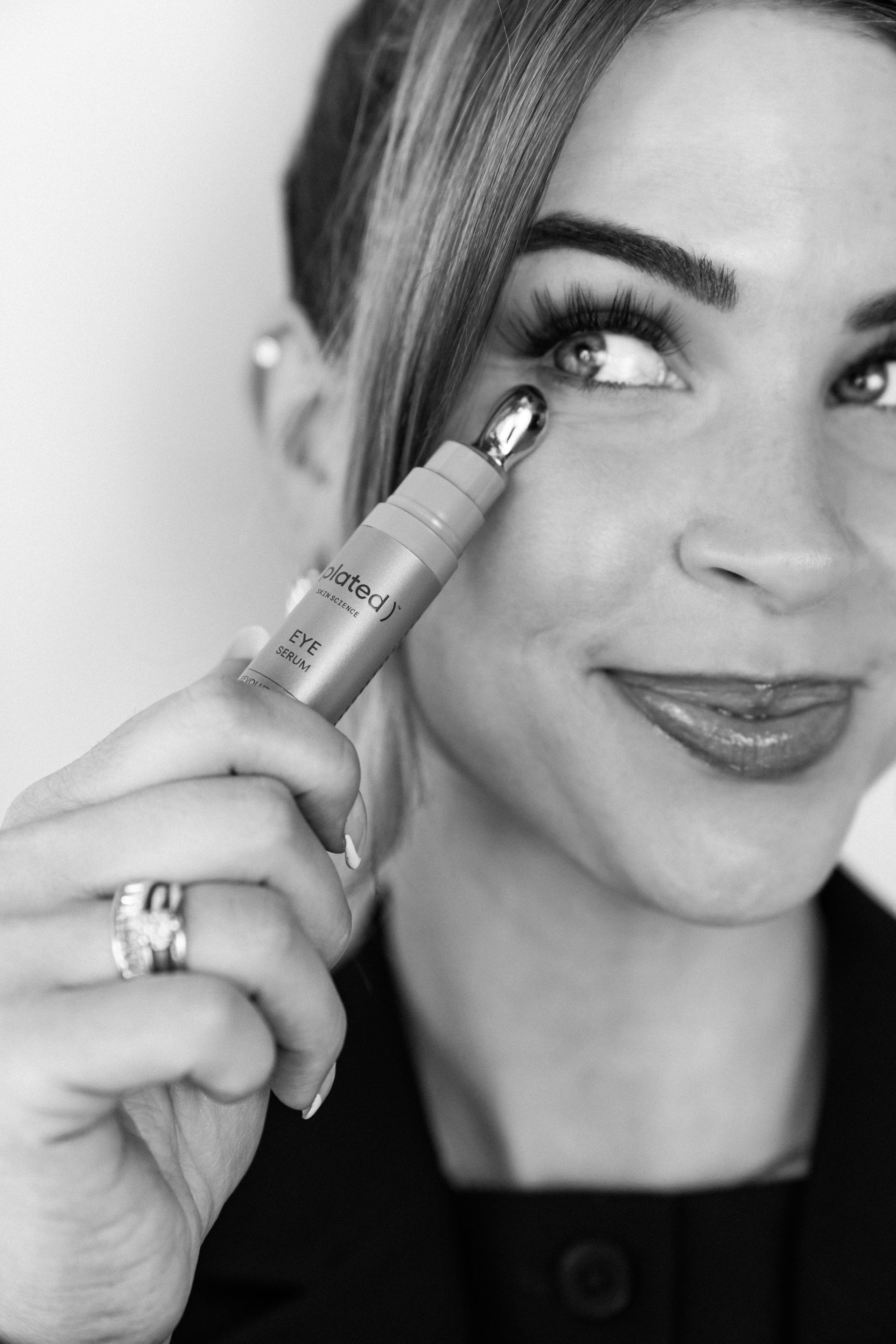 Woman applying eye makeup with a pencil, smiling in a close-up black-and-white portrait.