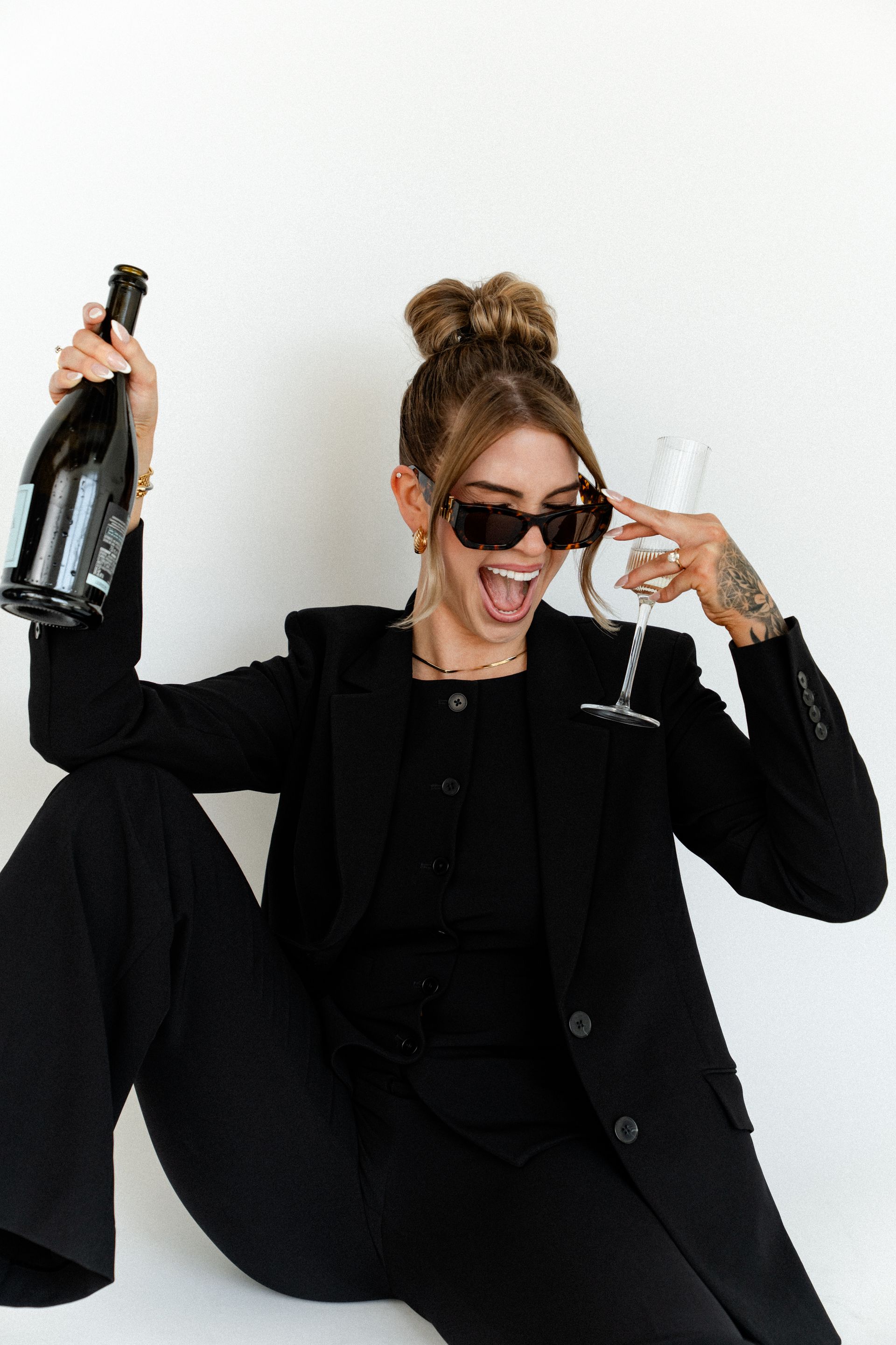 Woman in black suit and sunglasses holding a champagne bottle and glass, smiling on a white background