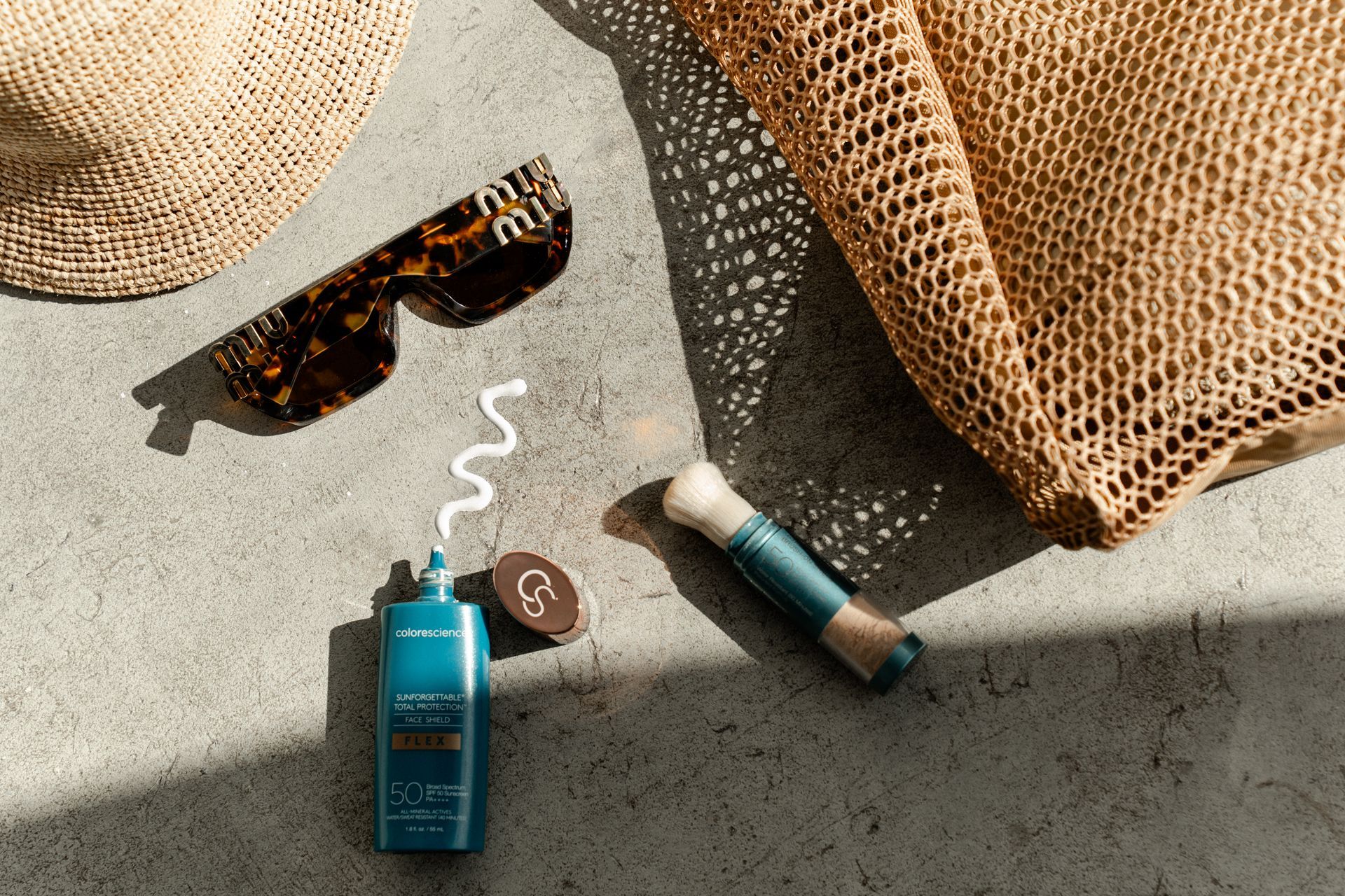 Sunglasses, sunscreen, and a vape beside woven beach bags on sunlit concrete