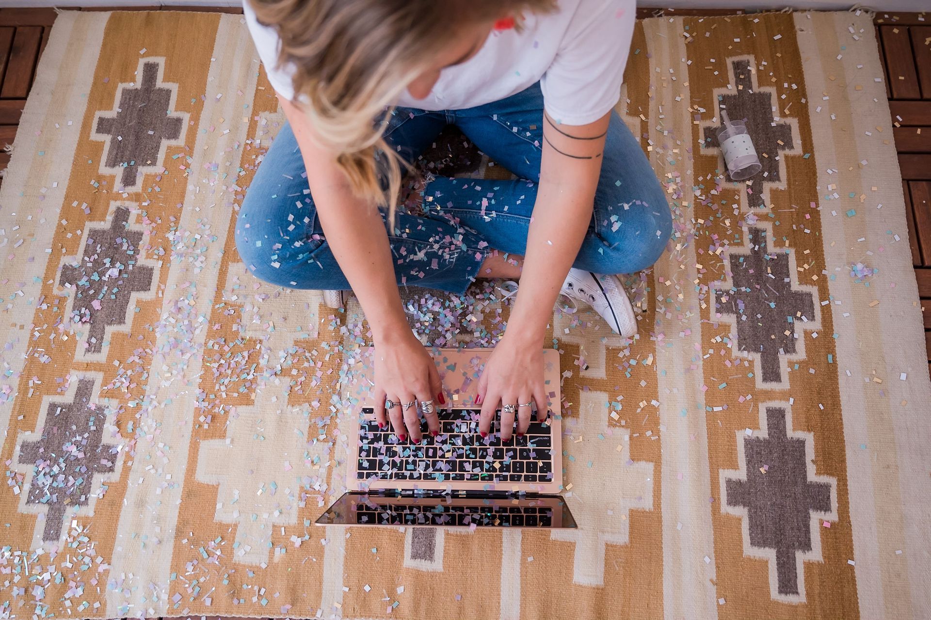 Person typing on a laptop on a patterned rug, viewed from above with glitter all around.