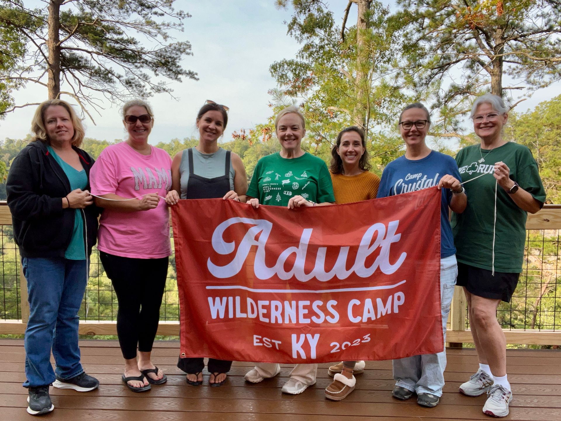 Seven people stand outdoors holding a red “Adult Wilderness Camp” banner in front of autumn trees.