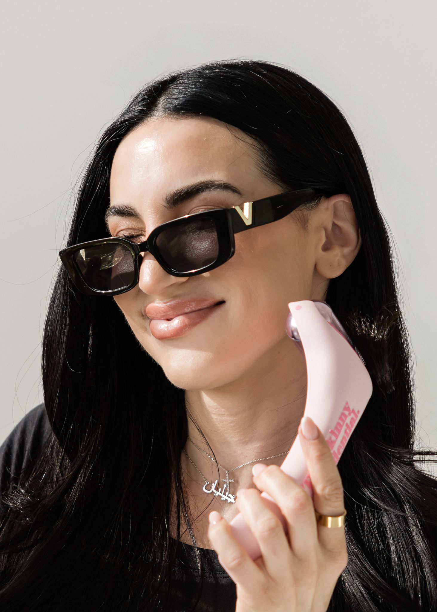 Woman wearing black sunglasses and holding a pink hairbrush, smiling against a light background