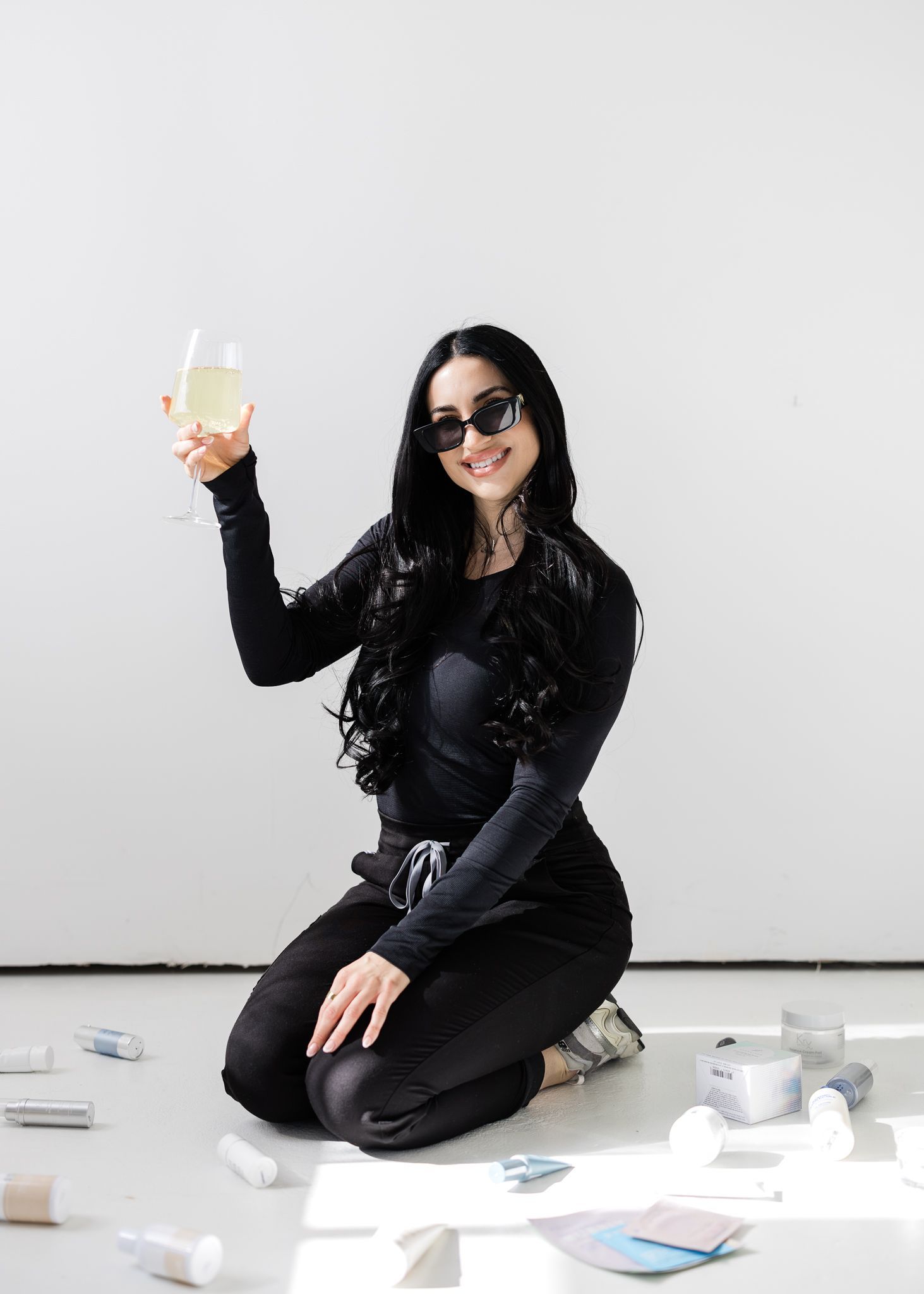 Woman in black kneels on a white floor, smiling and raising a drink against a plain white wall.