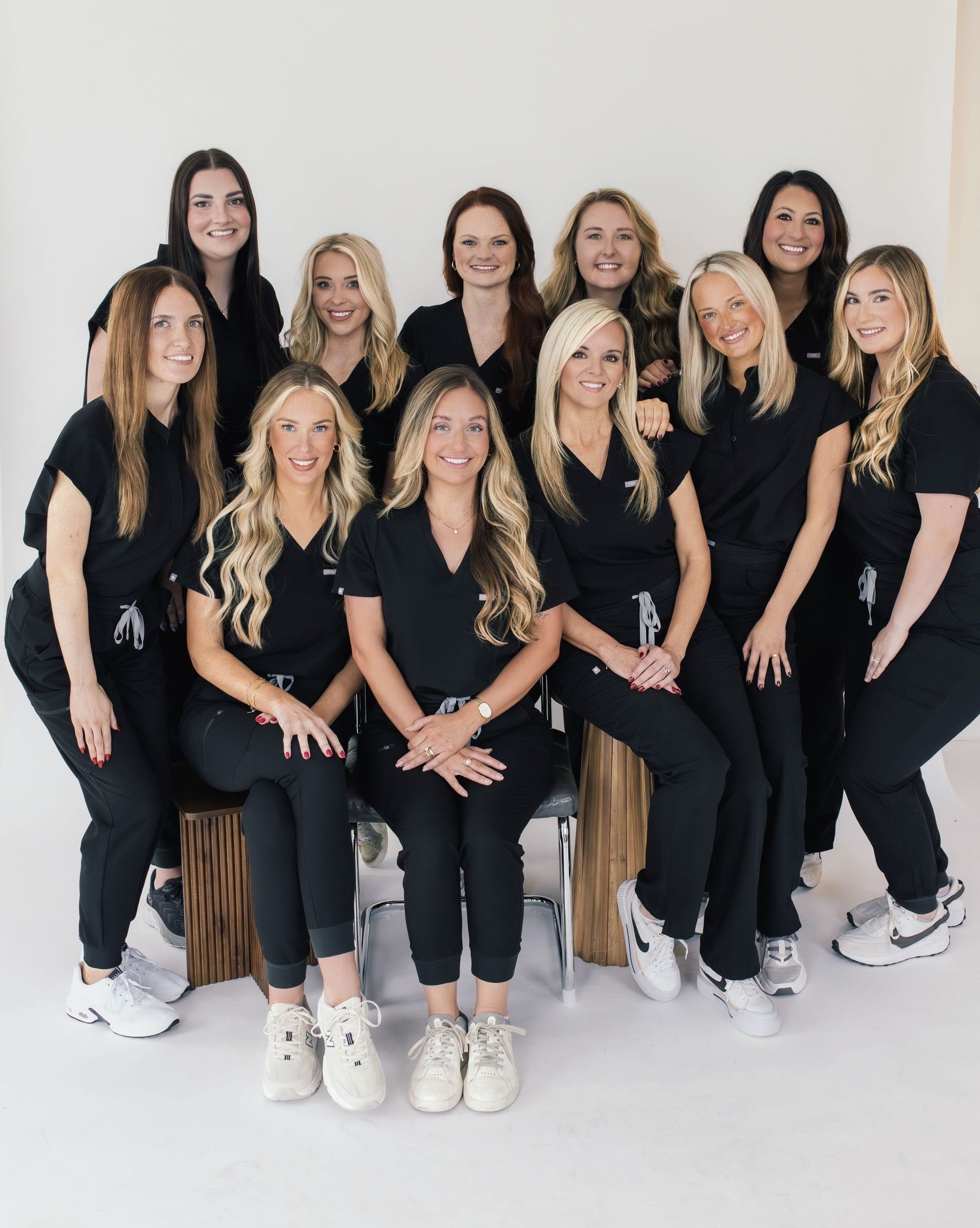 A group of eleven women in black scrubs and white sneakers posing for a photo in front of a white background.