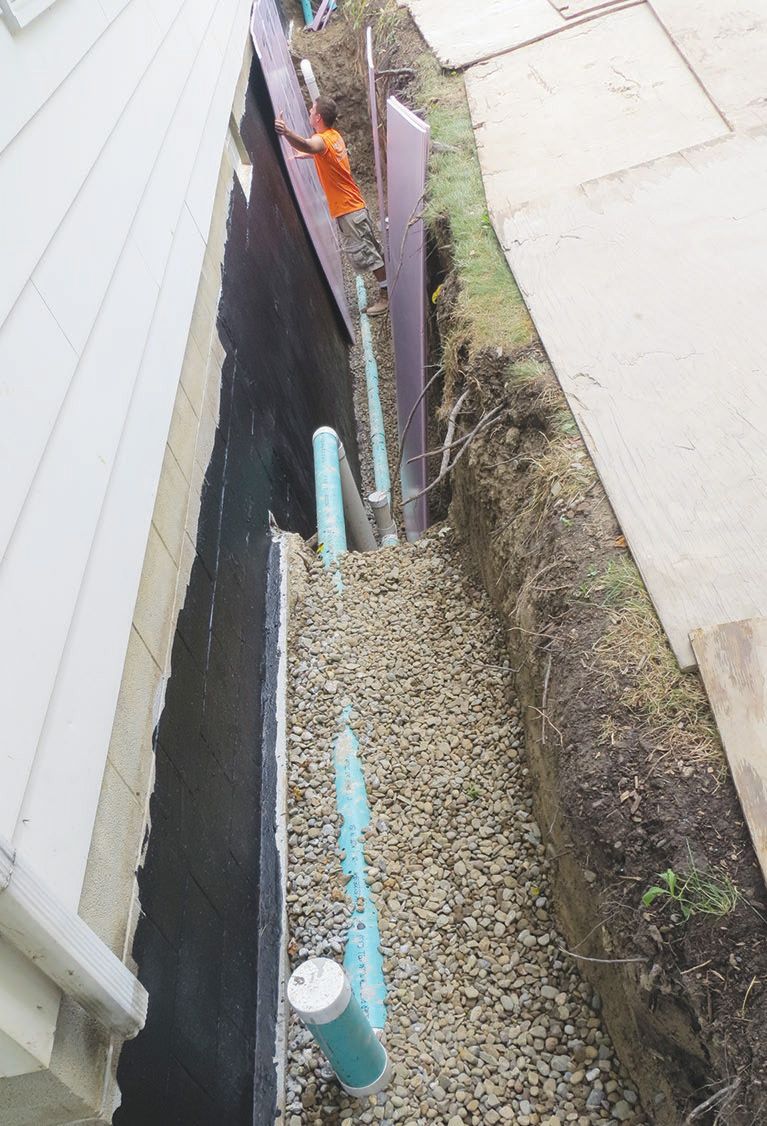 Construction of a narrow trench along a building's wall; gravel and pipes are visible. A person in orange shirt is working.