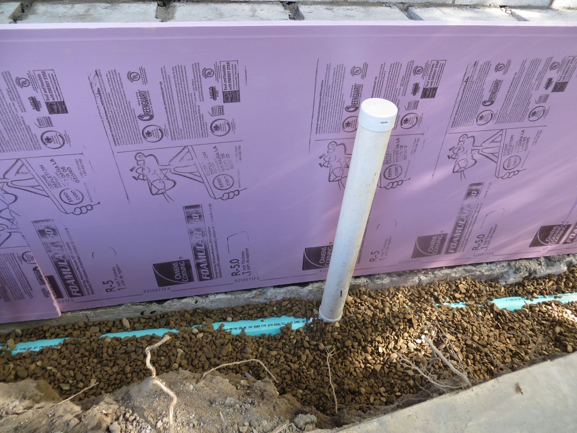 Pink insulation, white pipe, and gravel against a concrete foundation.