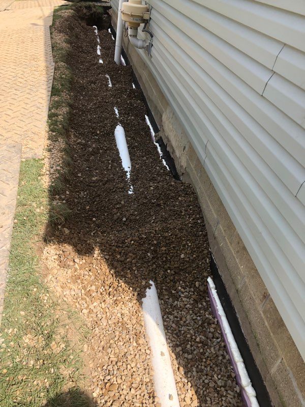 Gravel bed along house foundation with drainage pipes and white siding. Grass and paving are visible.