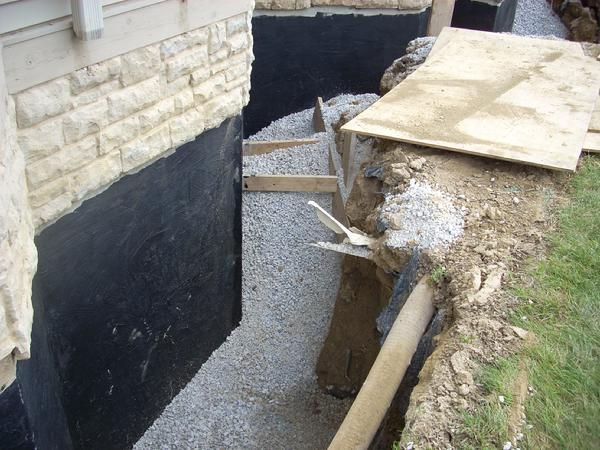 Foundation repair: gravel, black waterproofing, and a drainage pipe in a trench next to a building.