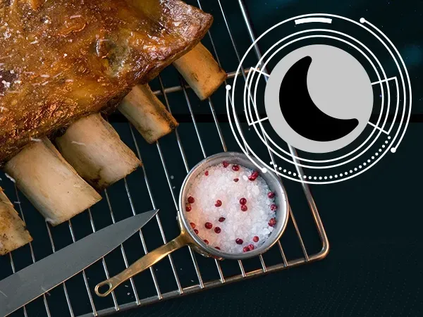 Un trozo de carne reposa sobre una rejilla junto a un bol de granos de pimienta rosa.